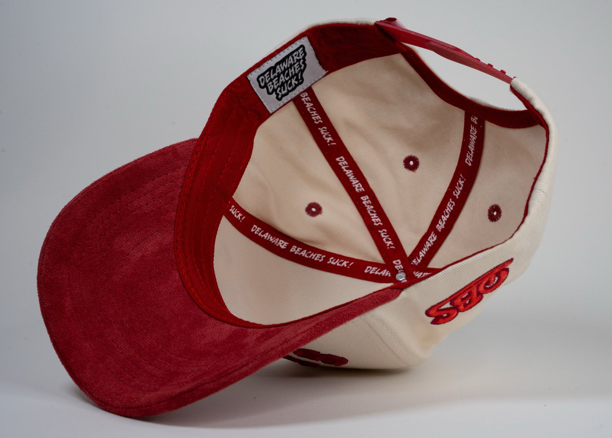 A cream baseball cap with red brim displays mirrored letters 'LEWES' and a red embroidered logo on the side.