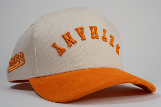 A cream baseball cap with an orange brim displays mirrored letters 'BETHANY' and an orange embroidered logo on the side.