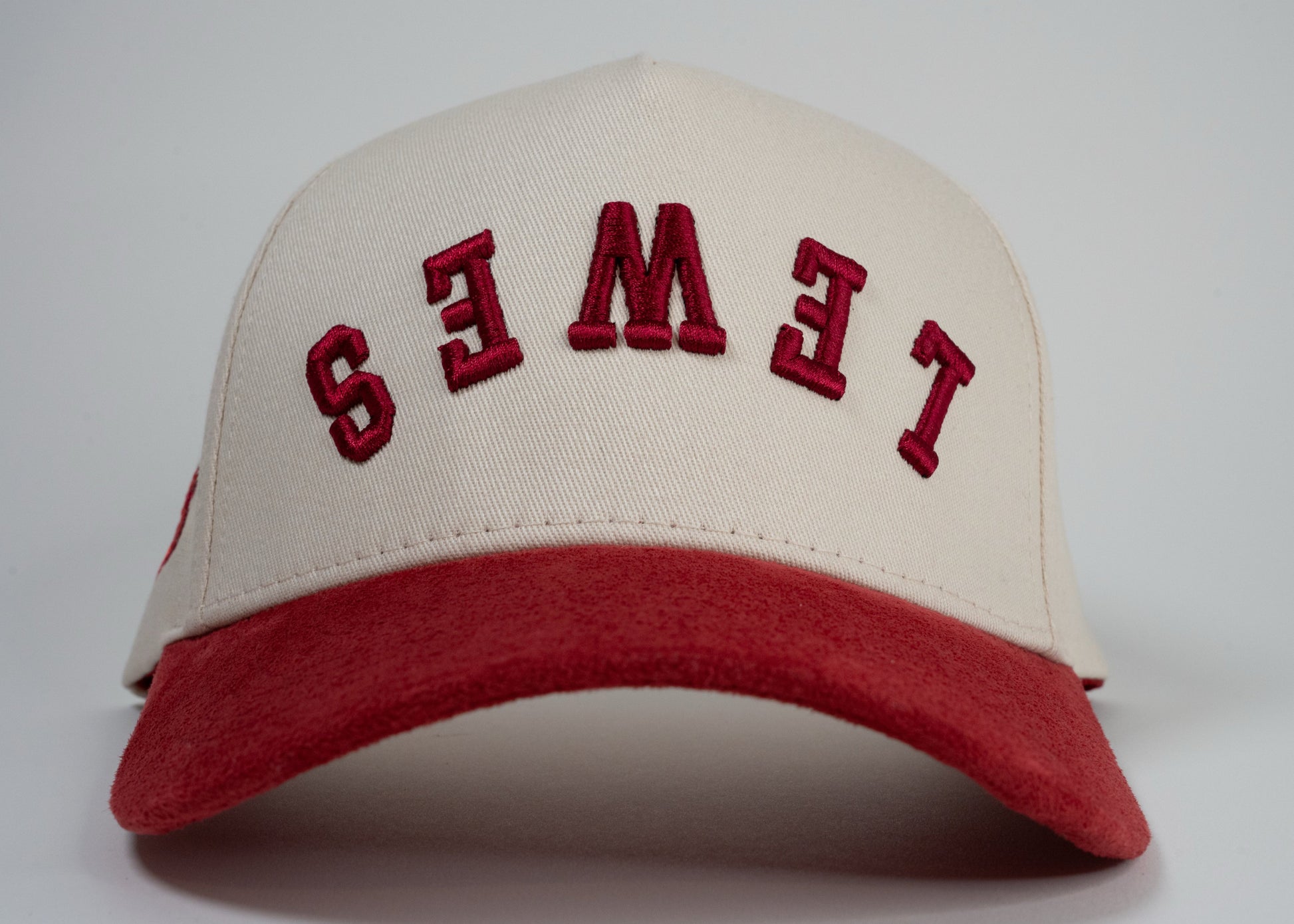 A cream baseball cap with red brim displays mirrored letters 'LEWES' and a red embroidered logo on the side.