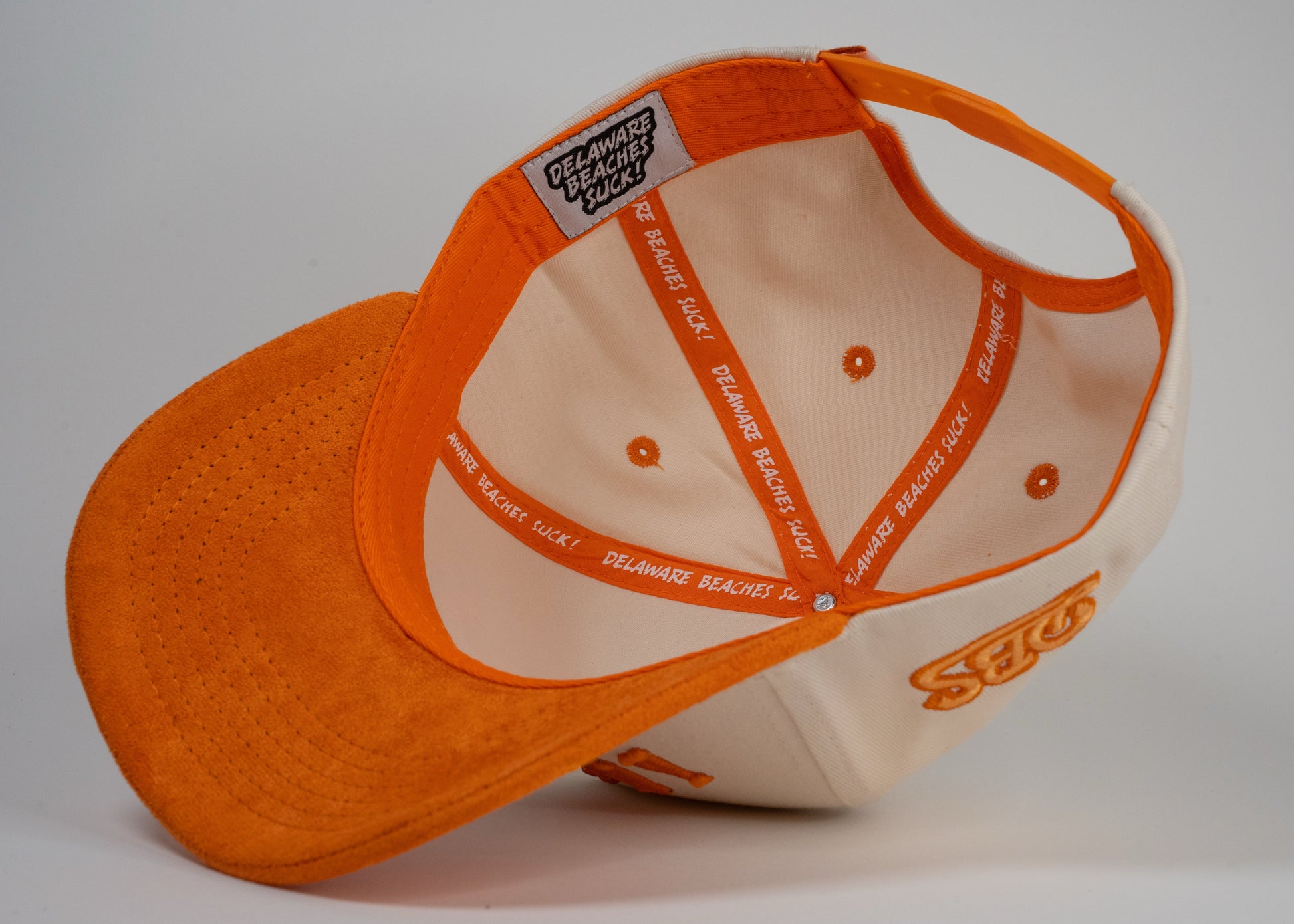 A cream baseball cap with an orange brim displays mirrored letters 'BETHANY' and an orange embroidered logo on the side.
