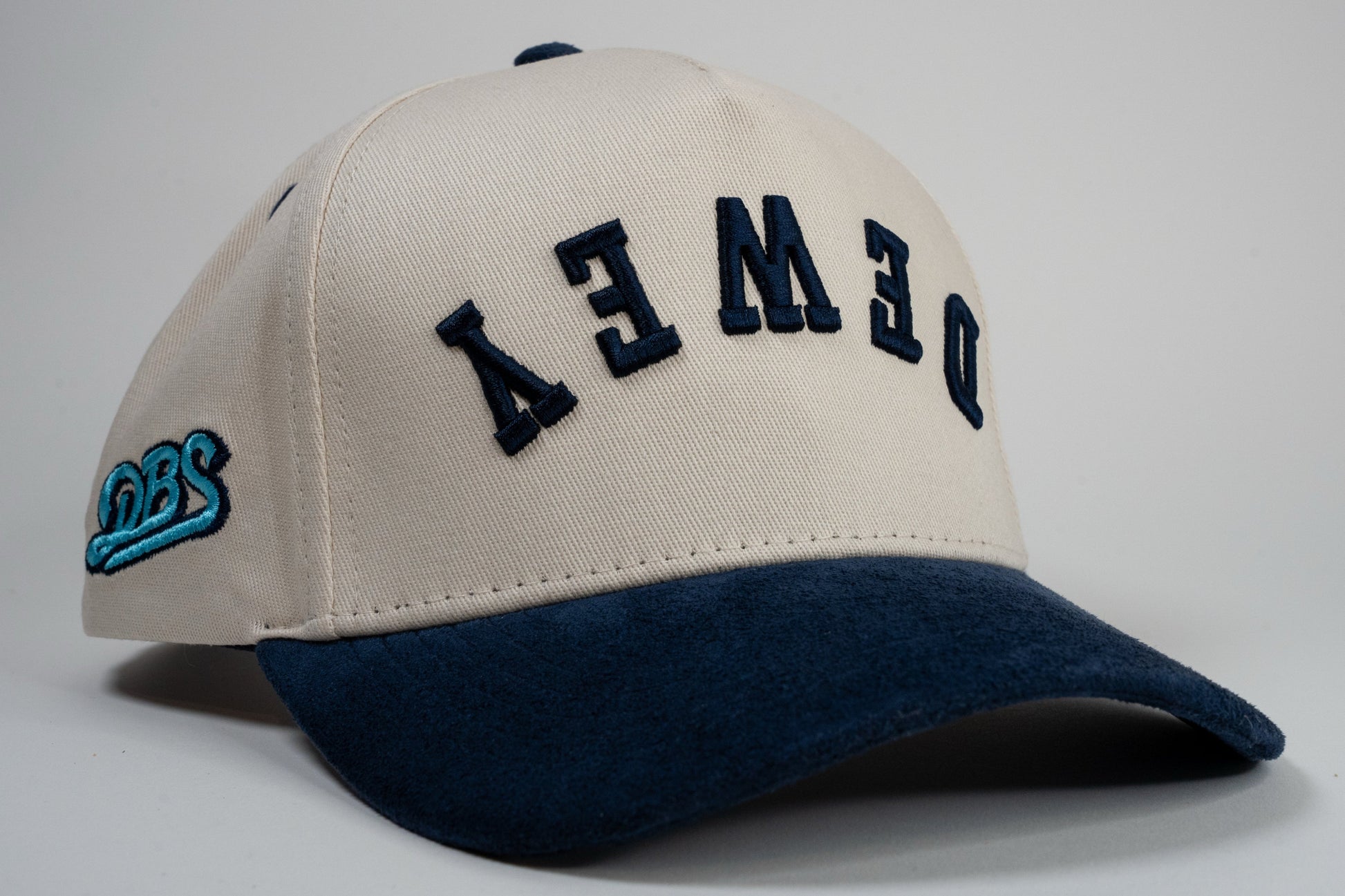 A cream baseball cap with navy blue brim displays mirrored letters 'DEWEY' and a blue embroidered logo on the side.