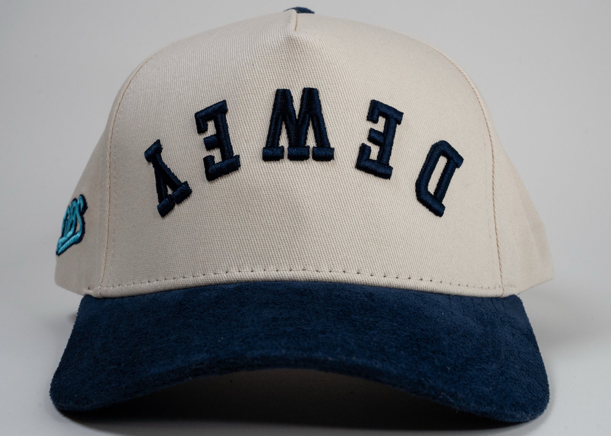 A cream baseball cap with navy blue brim displays mirrored letters 'DEWEY' and a blue embroidered logo on the side.
