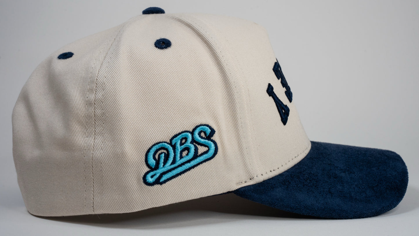 A cream baseball cap with navy blue brim displays mirrored letters 'DEWEY' and a blue embroidered logo on the side.