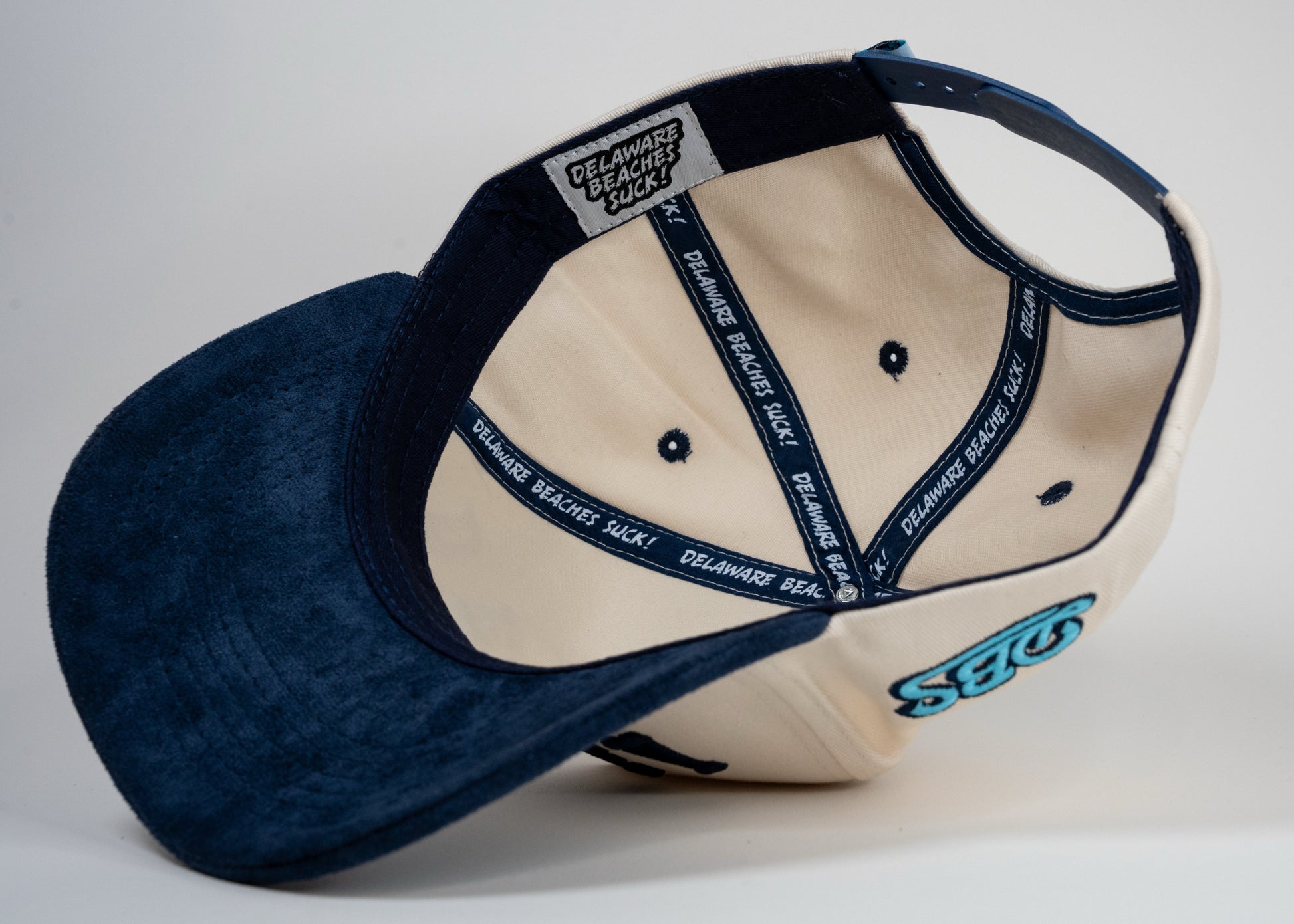 A cream baseball cap with navy blue brim displays mirrored letters 'DEWEY' and a blue embroidered logo on the side.