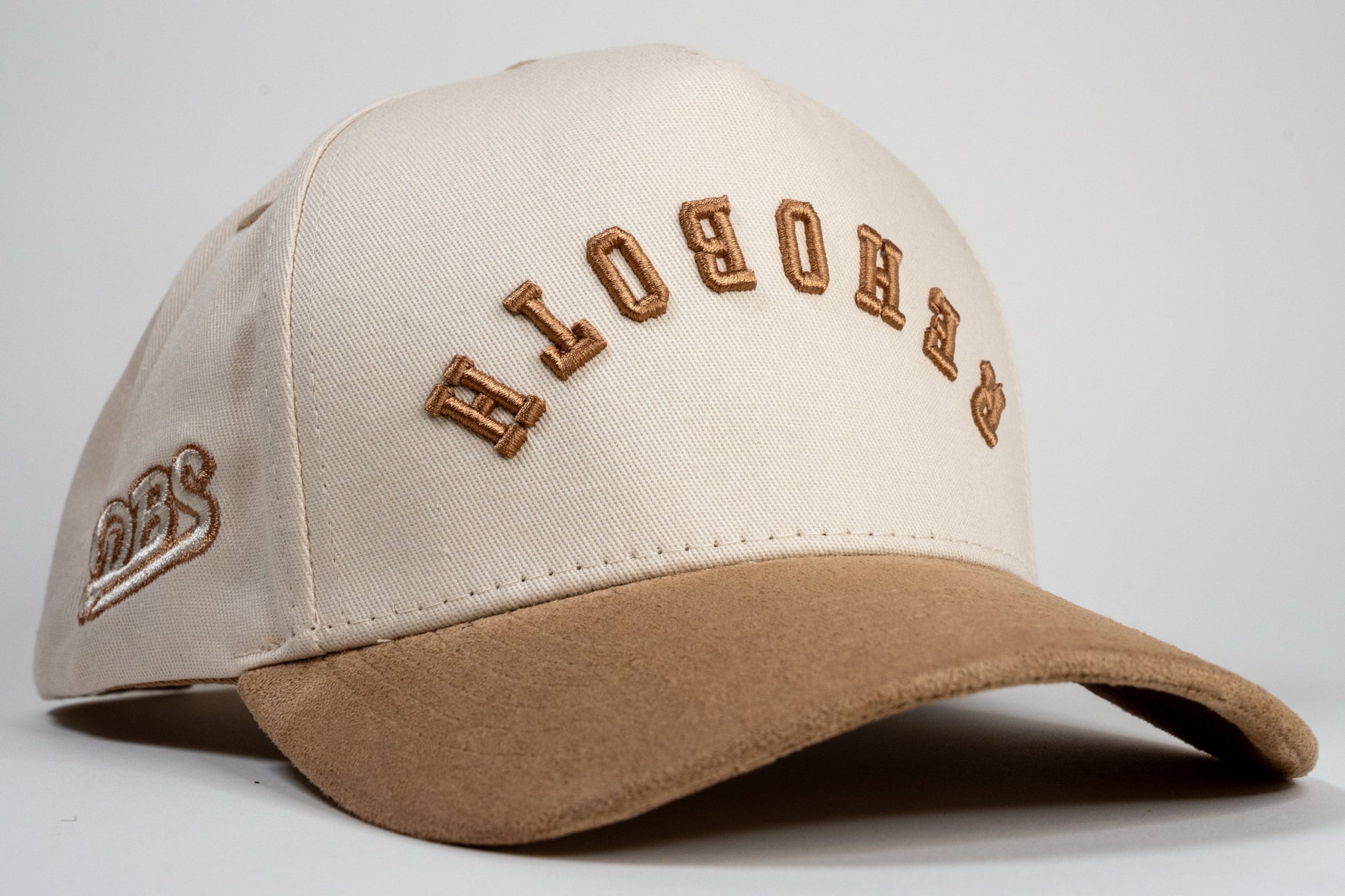 A cream baseball cap with beige brim displays mirrored letters 'REHOBOTH' and a beige embroidered logo on the side.