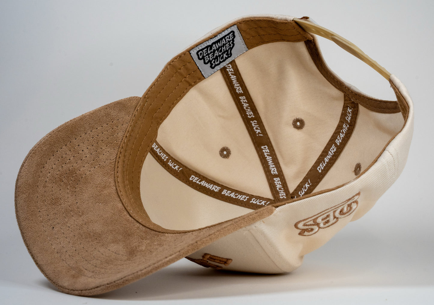 A cream baseball cap with beige brim displays mirrored letters 'REHOBOTH' and a beige embroidered logo on the side.