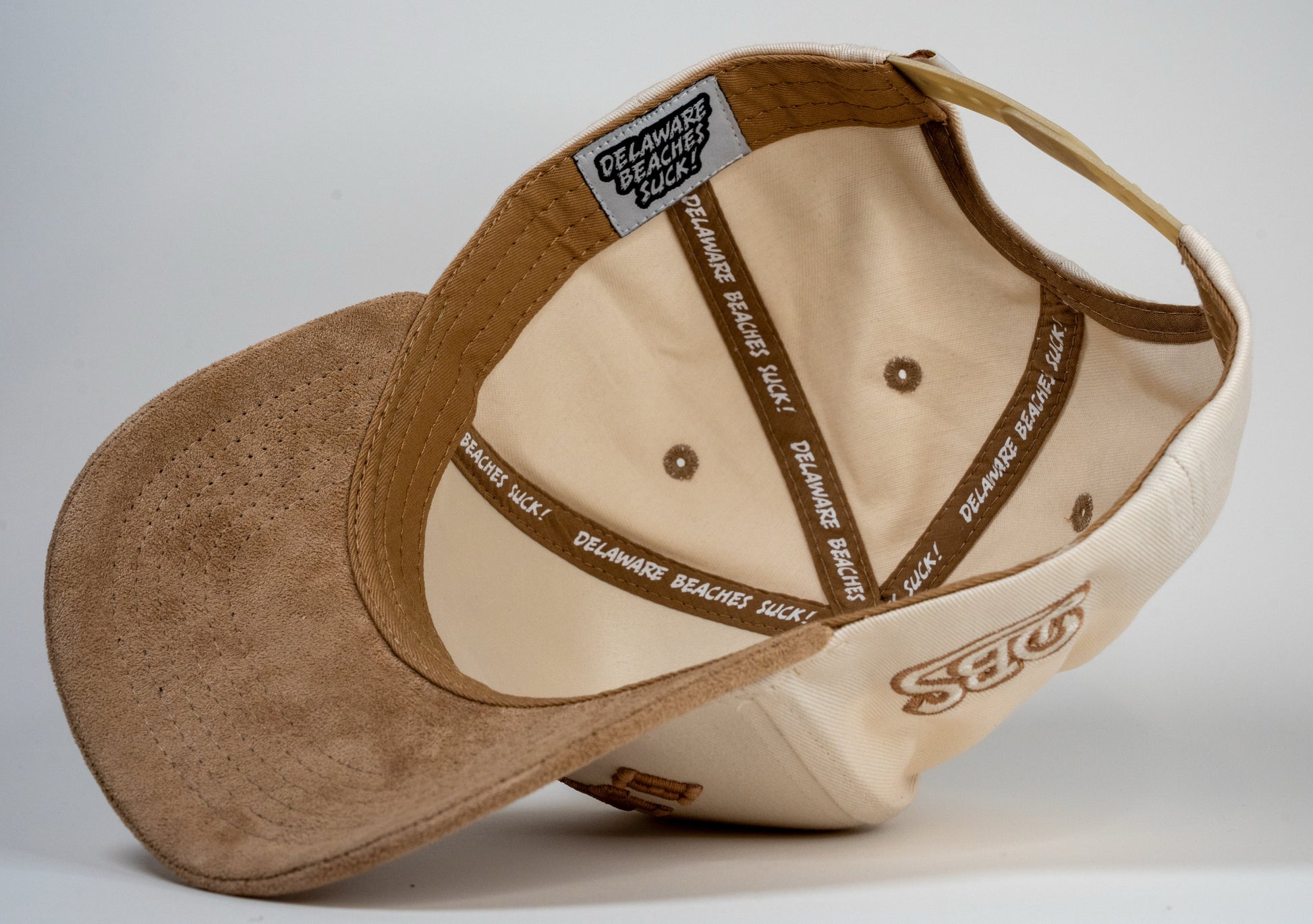A cream baseball cap with beige brim displays mirrored letters 'REHOBOTH' and a beige embroidered logo on the side.