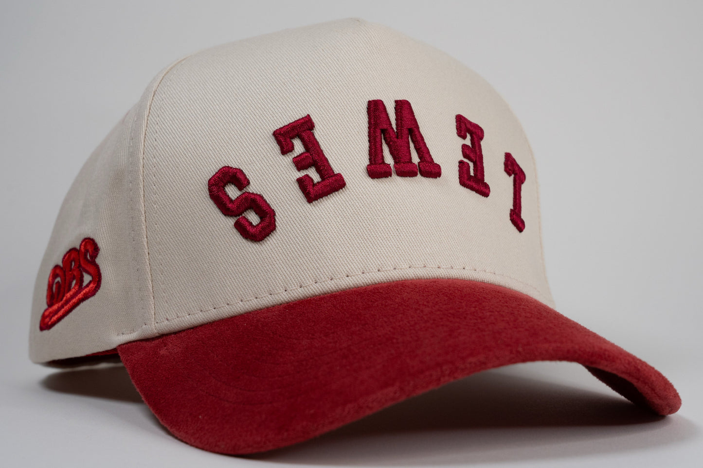 A cream baseball cap with red brim displays mirrored letters 'LEWES' and a red embroidered logo on the side.
