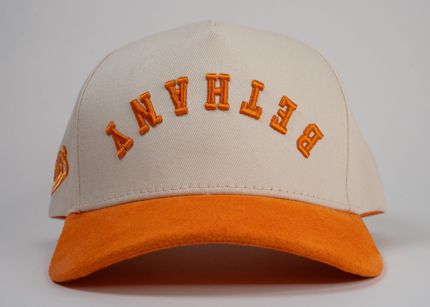 A cream baseball cap with an orange brim displays mirrored letters 'BETHANY' and an orange embroidered logo on the side.