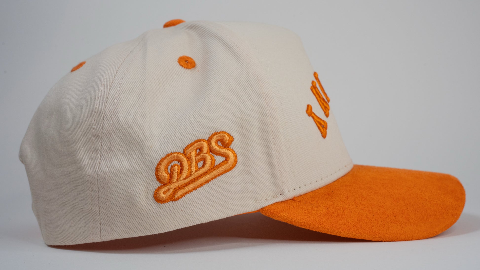 A cream baseball cap with an orange brim displays mirrored letters 'BETHANY' and an orange embroidered logo on the side.